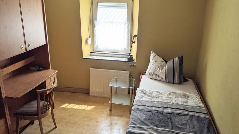 Ein einfaches Zimmer mit einem Bett, einem Tisch und einem Stuhl. Das Fenster lässt viel Tageslicht herein.
