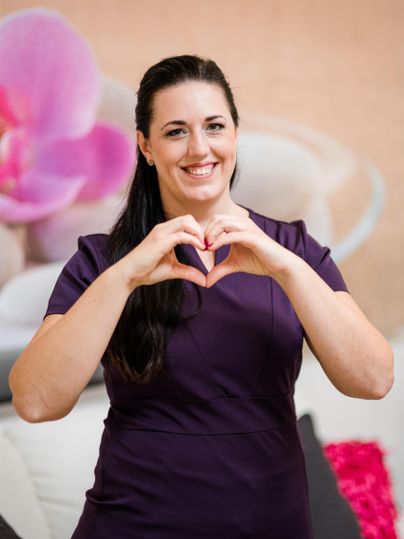 Eine lächelnde Frau in einem lila Kleid formt mit ihren Händen ein Herz. Im Hintergrund sind sanfte Farben und Blumen zu sehen.