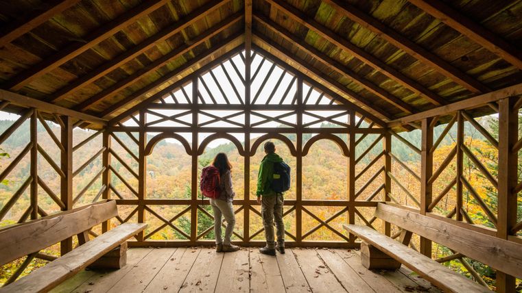 Zwei Wanderer stehen in einer Holzhütte mit großen Fenstern und bewundern die herbstliche Landschaft. Die Hütte ist einfach, aber einladend und bietet einen schönen Ausblick.