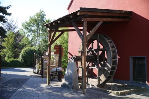 Ein historisches Wasserrad steht an einem Gebäude mit roter Fassade. Im Hintergrund sind Bäume zu sehen, die die Umgebung begrünen.