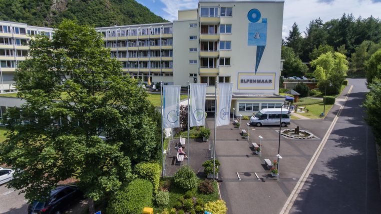 Ein modernes Gebäude mit einer großen Parkplatzfläche und umgebenden Bäumen. Der Himmel ist teilweise bewölkt und die Umgebung wirkt einladend.