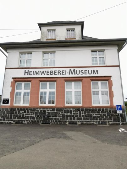 Ein Gebäude mit der Aufschrift "Heimweberei-Museum". Es hat mehrere Fenster und ist in einem historischen Stil erbaut.