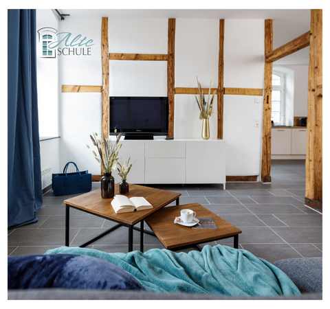 Ein modernes Wohnzimmer mit Holzbalken und einem Fernseher an der Wand. Auf dem Tisch liegen ein Buch und eine Tasse, ergänzt durch eine stilvolle Dekoration.