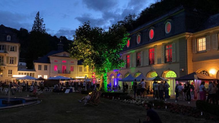 Eine festliche Atmosphäre bei Nacht mit beleuchtetem Gebäude und vielen Menschen im Freien. Bunte Lichter und eine grüne Platane schaffen eine lebendige Stimmung.