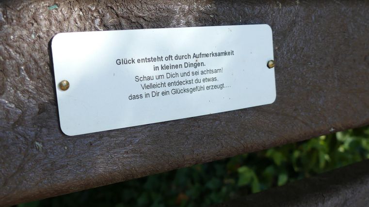 Eine Bank mit einer Metalltafel, auf der ein Zitat über das Glück steht. Im Hintergrund ist viel grün von Bäumen sichtbar.