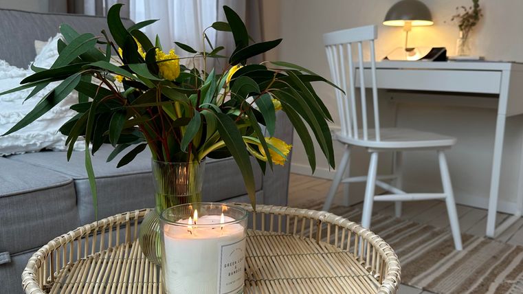 Ein gemütlicher Wohnraum mit einem runden Beistelltisch und einer Vase mit frischen Blumen. Im Hintergrund steht ein Schreibtisch mit einer Lampe und hellen Vorhängen.