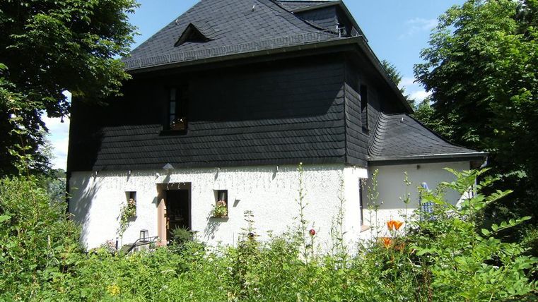 Ein charmantes, schwarzes Haus mit einem weißen Unterteil in einer grünen Umgebung. Umgeben von üppigen Pflanzen und Bäumen strahlt es eine ruhige Atmosphäre aus.