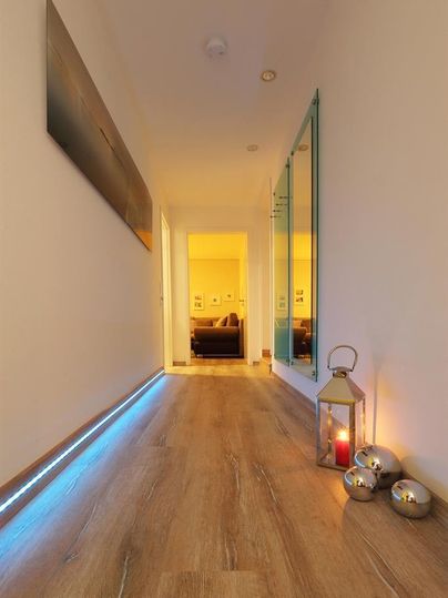 Ein heller Flur mit Holzfußboden und indirekter Beleuchtung. Am Ende des Flurs ist ein gemütliches Wohnzimmer sichtbar, und eine Laterne mit Kerze sorgt für eine einladende Atmosphäre.