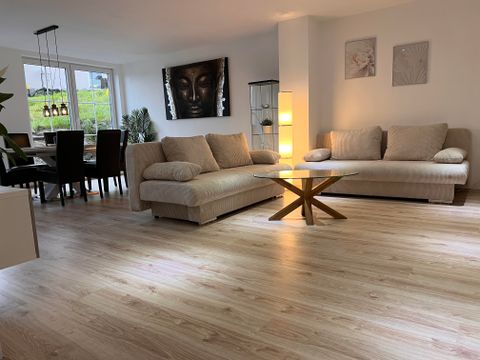 Ein modernes Wohnzimmer mit hellen Möbeln und einem großen Fenster. Es gibt eine gemütliche Sitzecke und einen Esstisch im Hintergrund.