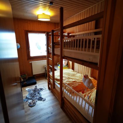 Ein gemütliches Zimmer mit einem Etagenbett und einem bunten Bettzeug. Der Raum hat holzverkleidete Wände und viel Tageslicht.