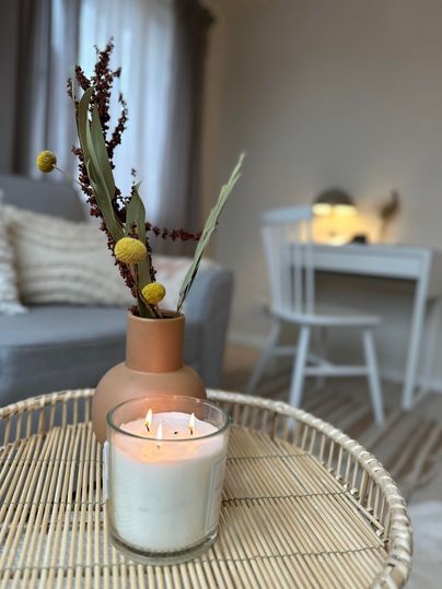 Ein gemütlicher Tisch mit einer Kerze und einem Blumengesteck in einer Vase. Im Hintergrund sind Möbel und ein neutraler Raum sichtbar.