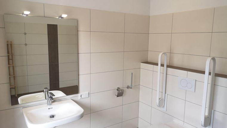 Ein modernes Bad mit hellem Fliesenboden, einem Waschbecken und einem Spiegel. An der Wand sind Haltegriffe für die Sicherheit angebracht.