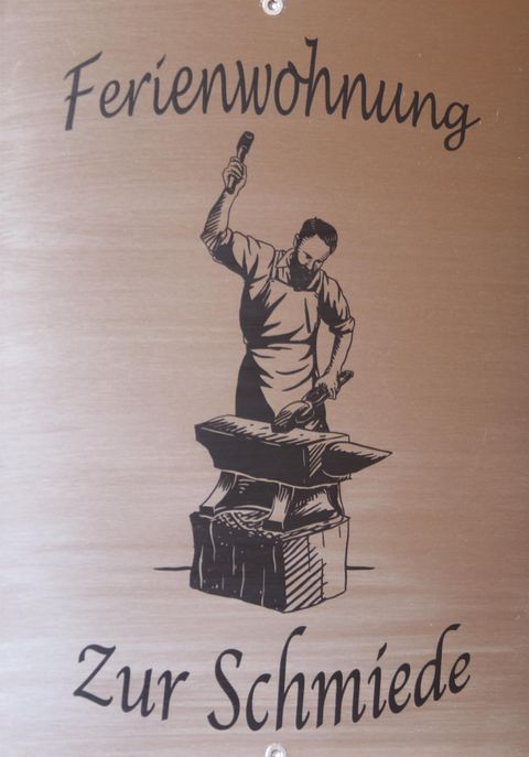 Eine Kunschrift mit der Aufschrift „Ferienwohnung Zur Schmiede“. Darauf ist eine stilisierte Darstellung eines Schmieds zu sehen, der an einemamboss arbeitet.