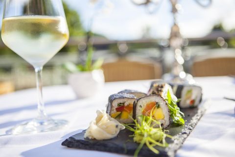 Ein eleganter Tisch mit Sushi-Rollen auf einem schwarzen Stein. Im Hintergrund steht ein Glas Weißwein und eine kleine Blume.