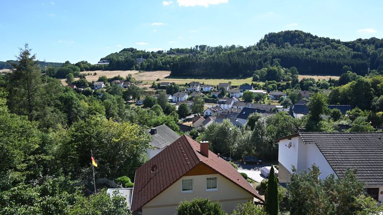 Eine malerische Landschaft mit vielen Häusern und üppigem Grün. Im Hintergrund sind sanfte Hügel und ein klarer blauer Himmel zu sehen.