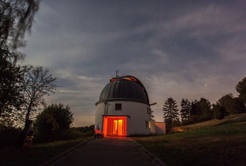 Eine Astronomieteleskop-Observatorium bei Nacht, beleuchtet von rotem Licht. Umgeben von Bäumen und einem klaren Himmel.