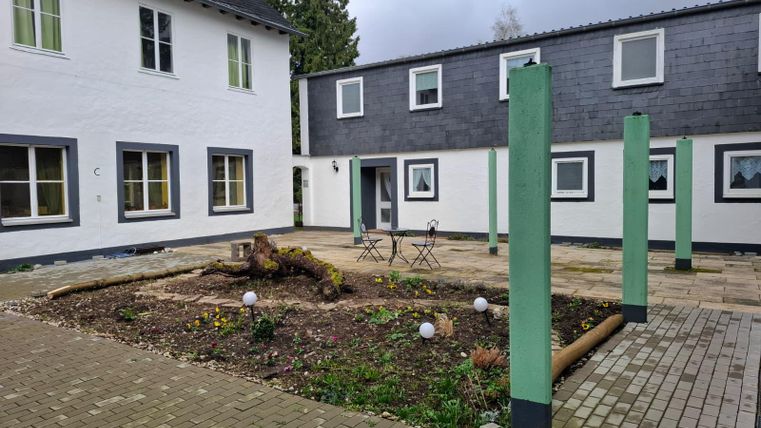 Ein kleiner Innenhof mit zwei Gebäuden und einer begrünten Fläche in der Mitte. Es gibt einige Stühle und Säulen sowie dekorative Kugeln.