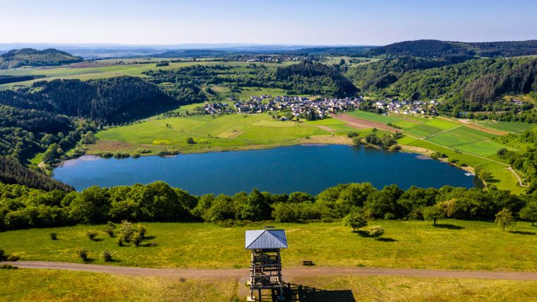 Luftaufnahme des Meerfelder Maars mit einem Aussichtsturm im Vordergrund und einem Dorf im Hintergrund.