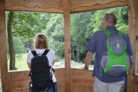 Zwei Personen stehen in einer Holzhütte und schauen nach draußen auf eine grüne Landschaft. Beide tragen Rucksäcke und genießen die Aussicht.