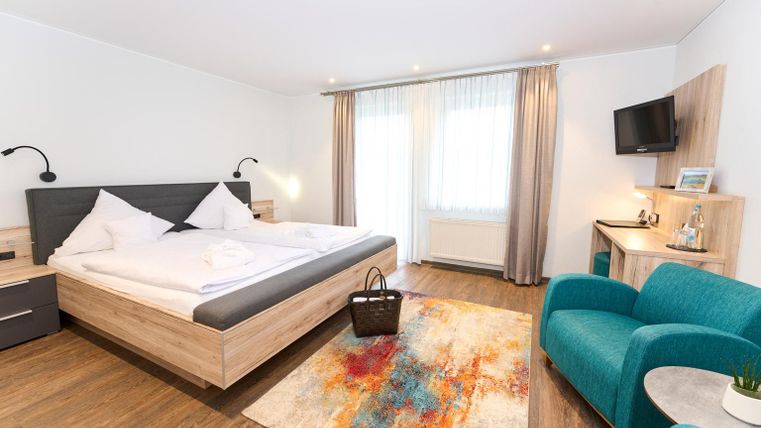 Ein modernes Gästezimmer mit einem großen Bett und einem gemütlichen Sessel. Der Raum ist hell und freundlich, mit einem bunten Teppich und einem Fernseher.