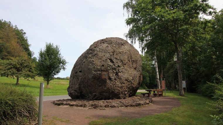 Ein großer, runder Felsen steht in einer grünen Umgebung. Daneben gibt es eine Bank und Bäume im Hintergrund.