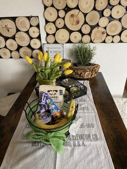 Ein Tisch mit einer Dekoration aus gelben Tulpen und einem Korb voller Snacks. Im Hintergrund sind Holzscheiben an der Wand zu sehen.