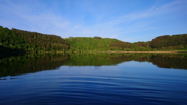Ein ruhiger See, umgeben von grünen Hügeln und Wäldern. Das Wasser spiegelt den klaren blauen Himmel wider.