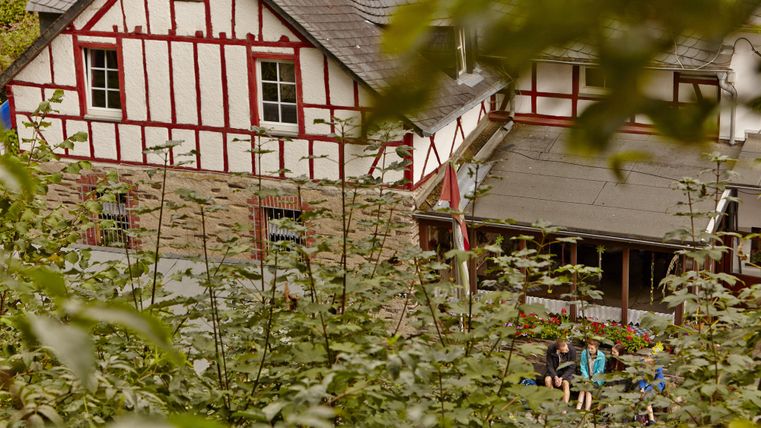 Ein historisches Gebäude mit rotem Fachwerk in einer grünen Umgebung. Im Vordergrund sind Personen, die entspannen und die Natur genießen.