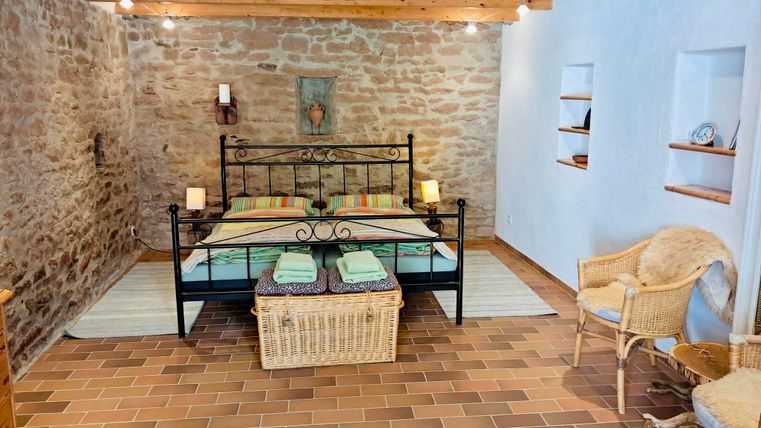 Ein gemütliches Schlafzimmer mit einem Doppelbett und rustikalem Steinmauerwerk. Die Einrichtung umfasst einen Korbsessel und offene Regale.