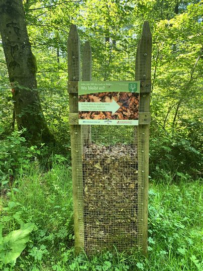 Ein Gitterbehälter im Wald, gefüllt mit Laub, mit einem Schild darüber.