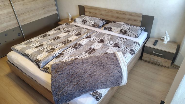 Ein einfaches Schlafzimmer mit einem Doppelbett, das mit einem Musterbettzeug bedeckt ist. Neben dem Bett steht ein Nachttisch mit einer kleinen Figur.