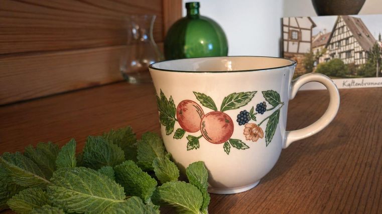 Eine Tasse mit einem bunten Obstmuster steht auf einem Holztisch. Neben der Tasse liegt frisches Minzblatt.