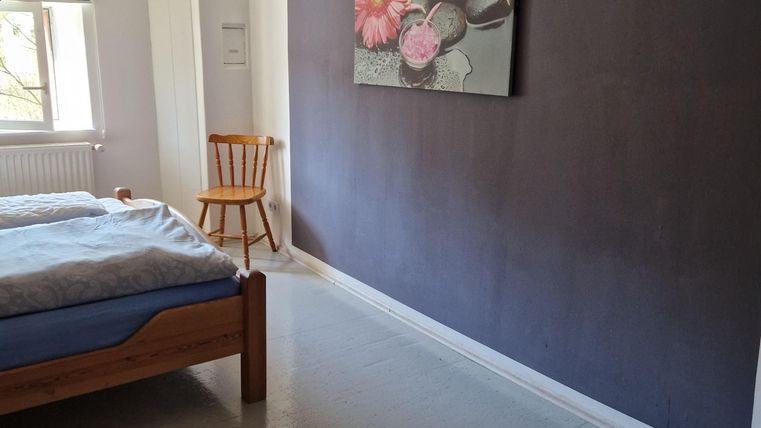 Ein einfaches Schlafzimmer mit einem Holzbett und einem Stuhl. An der Wand hängt ein gemaltes Bild von Blumen.