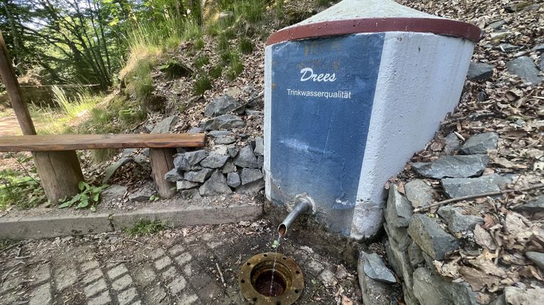 Ein Wasserhäuschen mit einer Quelle in der Natur. Daneben befindet sich eine Bank aus Holz.