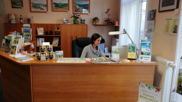 Ein gemütliches Büro mit einem Holzschreibtisch und einer Frau, die am Computer arbeitet. An den Wänden hängen Landschaftsbilder, und es gibt zahlreiche Informationsmaterialien auf dem Tisch.