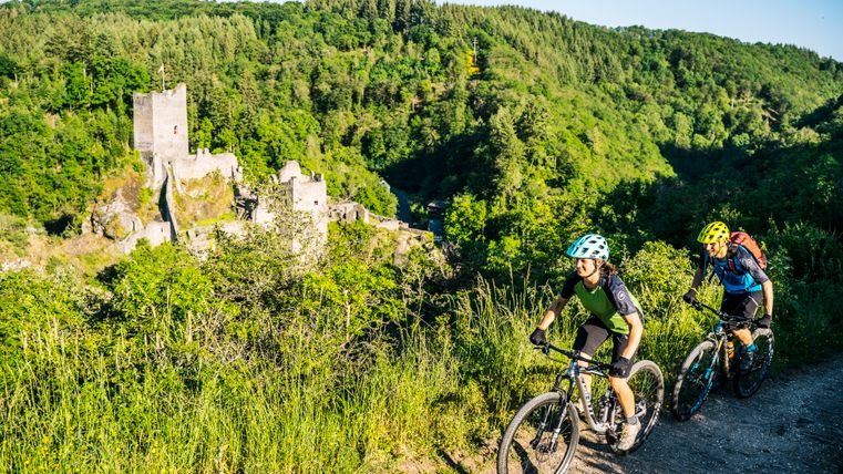 Zwei Mountainbiker fahren auf einem Pfad mit einer Burgruine im Hintergrund.