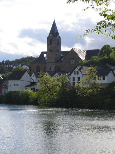 Eine malerische Kirche mit einem hohen Turm steht am Ufer eines ruhigen Sees. Umgeben von grünen Bäumen und bunten Gebäuden zeigt die Szene eine friedliche Landschaft.