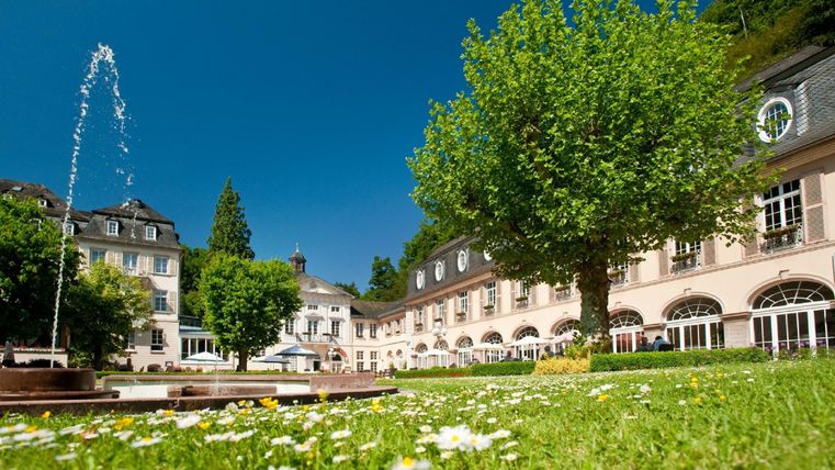 Ein schöner Park mit einem Brunnen und einer grünen Wiese voller Blumen. Im Hintergrund sind elegante Gebäude unter einem klaren blauen Himmel zu sehen.