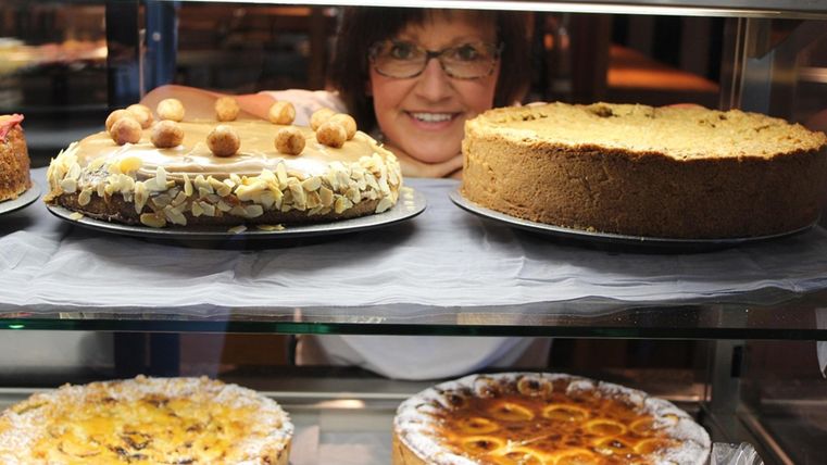 Eine freundliche Bäckerin präsentiert eine Auswahl an verschiedenen Kuchen in einer Vitrine. Die Kuchen sind reich verziert und appetitlich.