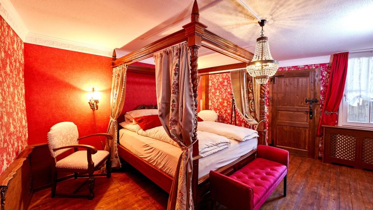 Ein gemütliches Schlafzimmer mit einem Himmelbett und roter Tapete. Der Raum ist stilvoll eingerichtet mit einem Sessel und einem Kristalllüster.
