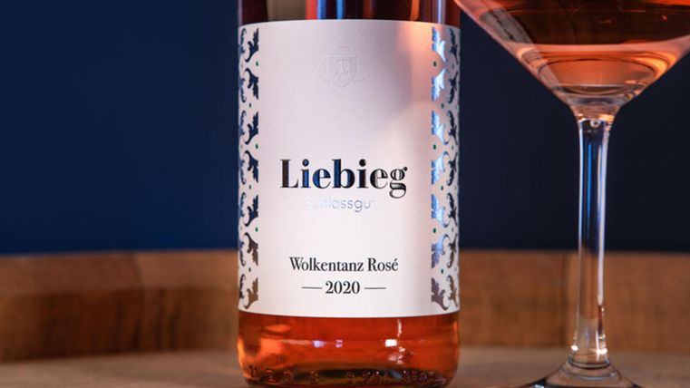 Eine Flasche Liebieg Wolkentanz Rosé 2020 steht auf einem Holztisch. Daneben befindet sich ein gefülltes Weinglas.