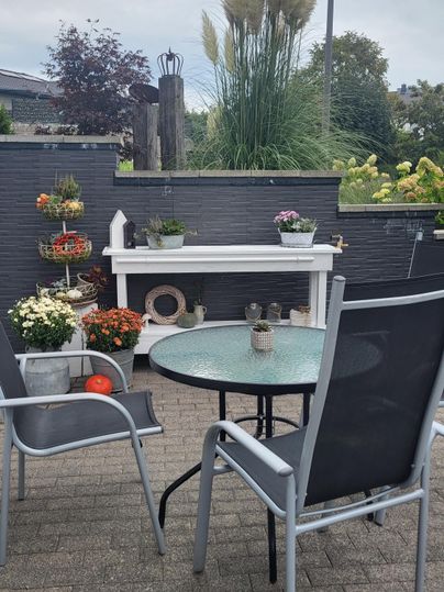 Eine gemütliche Terrasse mit einem runden Tisch und Stühlen. Bunte Blumen in Töpfen sorgen für eine einladende Atmosphäre.