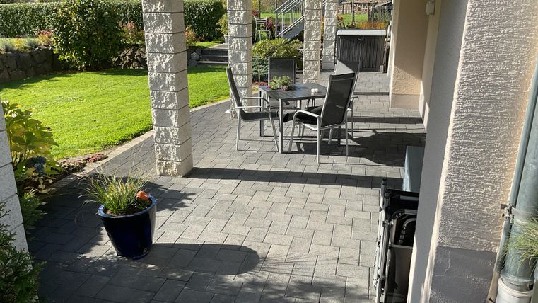 Eine gemütliche Terrasse mit Stühlen und einem Tisch, umgeben von grünem Rasen und Pflanzen. Die Sonne scheint auf den gepflasterten Boden.