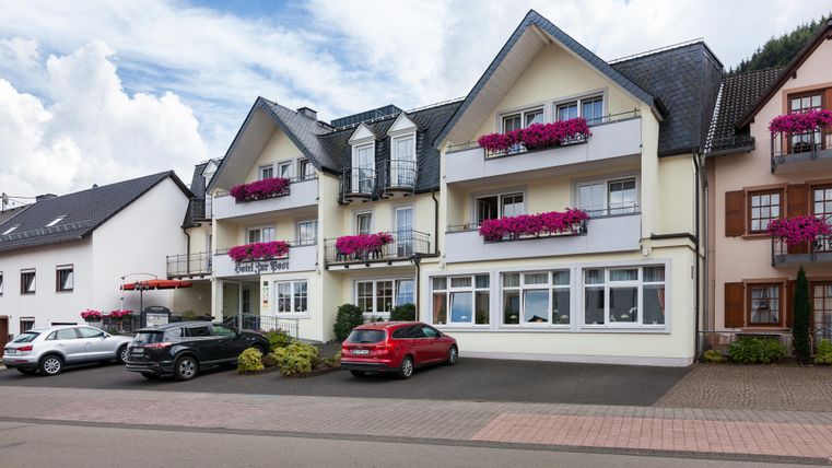 Ein gemütliches Wohnhaus mit Balkonen und bunten Blumenkästen. Dazu eine gepflasterte Straße mit mehreren parkenden Autos.