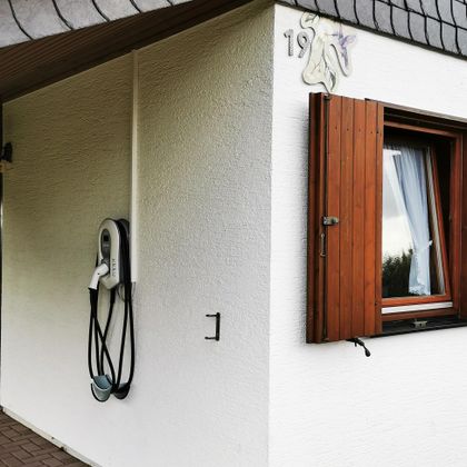 Eine Wand mit einem Elektroauto-Ladegerät und einem offenen Fenster. Die Fassade ist weiß mit einem Holzfenster.