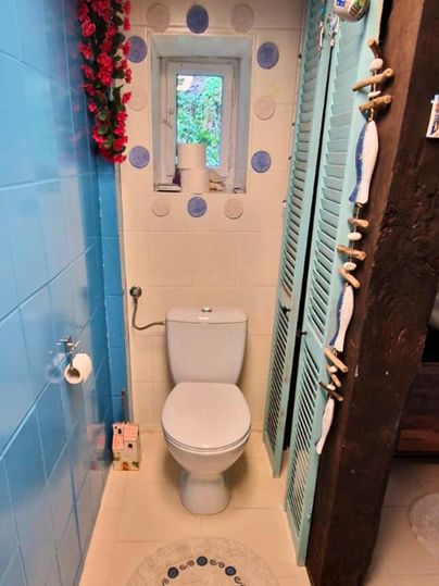 Ein helles Badezimmer mit blauen Fliesen und einem weißen WC. An der Wand sind dekorative Elemente und ein kleines Fenster mit Blick ins Freie.