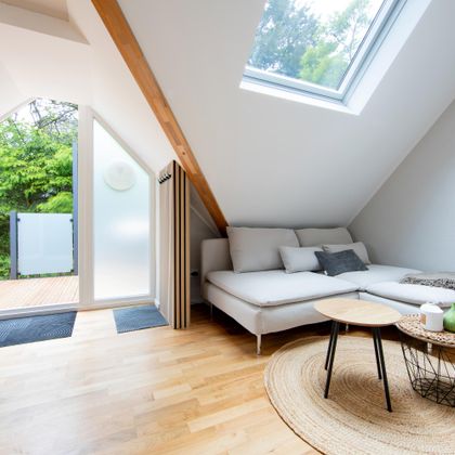 Ein modernes, lichtdurchflutetes Zimmer mit einem gemütlichen Sofa und einem kleinen Tisch. Große Fenster bieten Aussicht auf einen grünen Außenbereich.