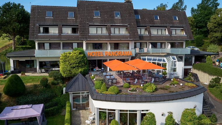 Ein modernes Hotel mit der Aufschrift "Hotel Panorama". Es hat eine Terrasse mit orangefarbenen Sonnenschirmen und ist von gepflegten Gärten umgeben.