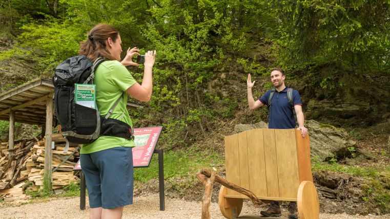A woman takes a photo of a man standing behind a wooden wagon on a hiking trail in the forest.