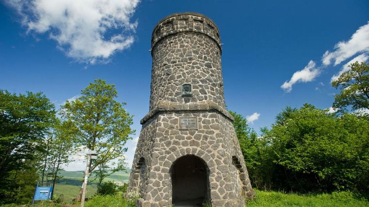 Ein historischer Steinturm steht umgeben von üppigem Grün und blauem Himmel. Die Sonne scheint, und die Wolken sind teilweise sichtbar.